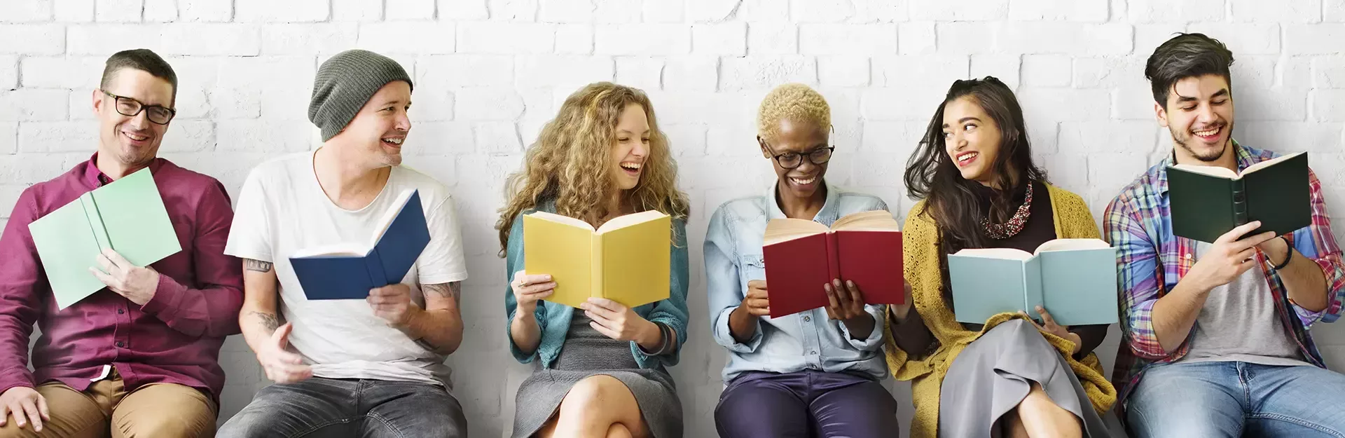 Studierende sitzen aufgereiht an der Wand und stöbern in Büchern 